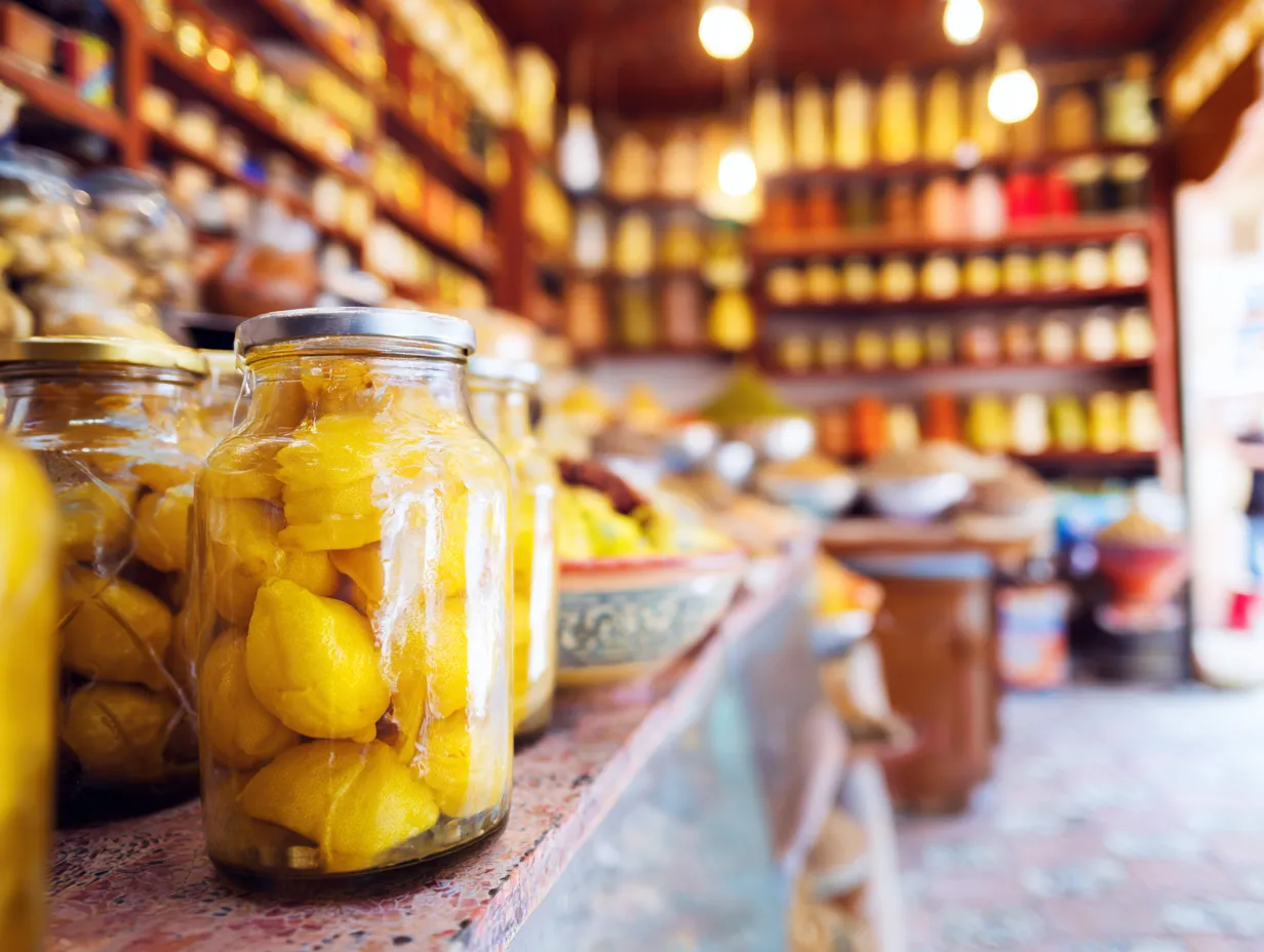 Moroccan Beldi Preserved Lemons: Ultimate Tangy Delight 11 Several glass jars of golden Moroccan preserved lemons displayed in a specialty food market, surrounded by colorful spices and exotic ingredients, ready for purchase.