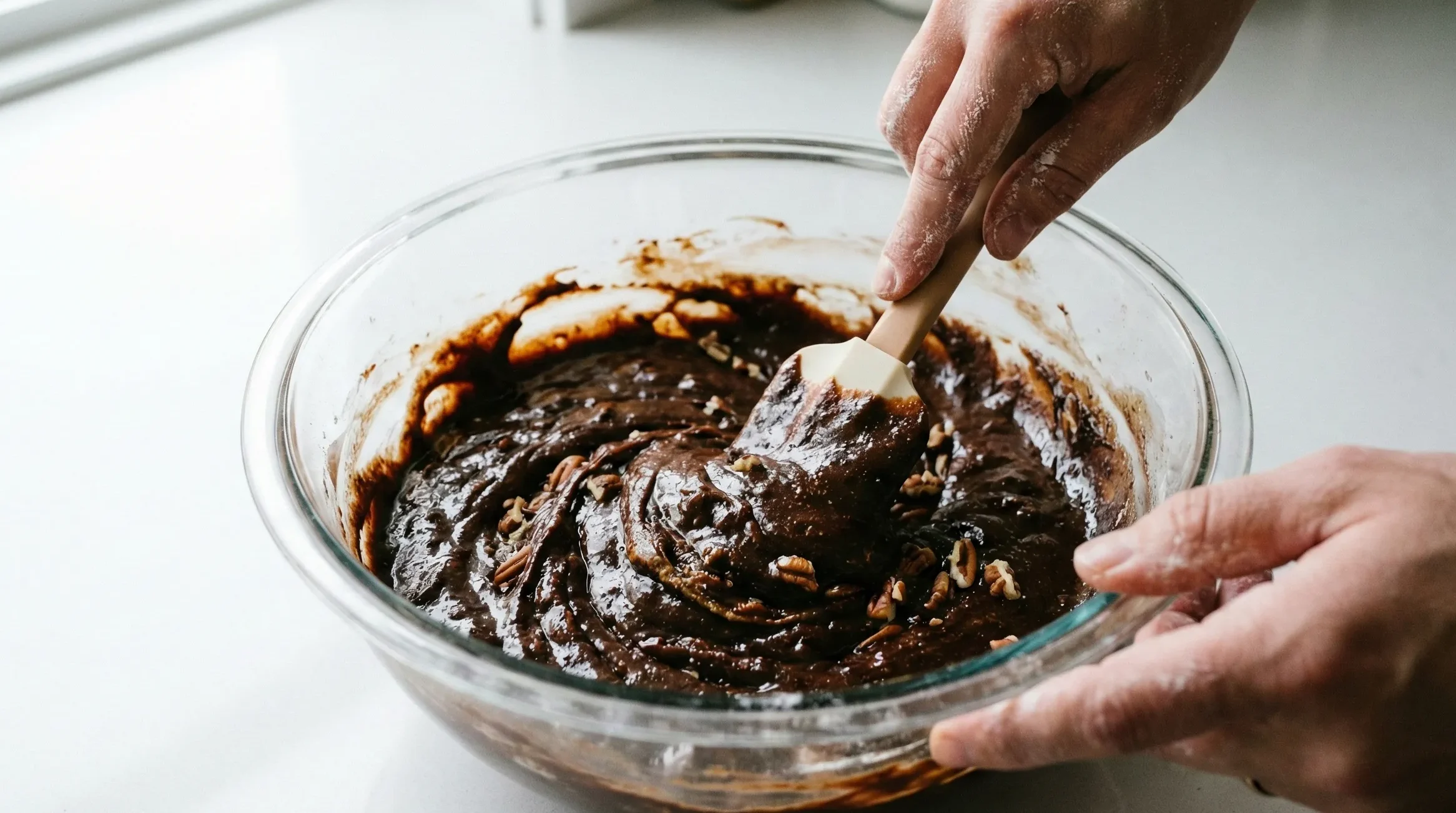 Close-up of hands gently folding the thick, glossy fudgy pecan date brownie batter in a glass bowl, demonstrating the crucial step of avoiding overmixing.